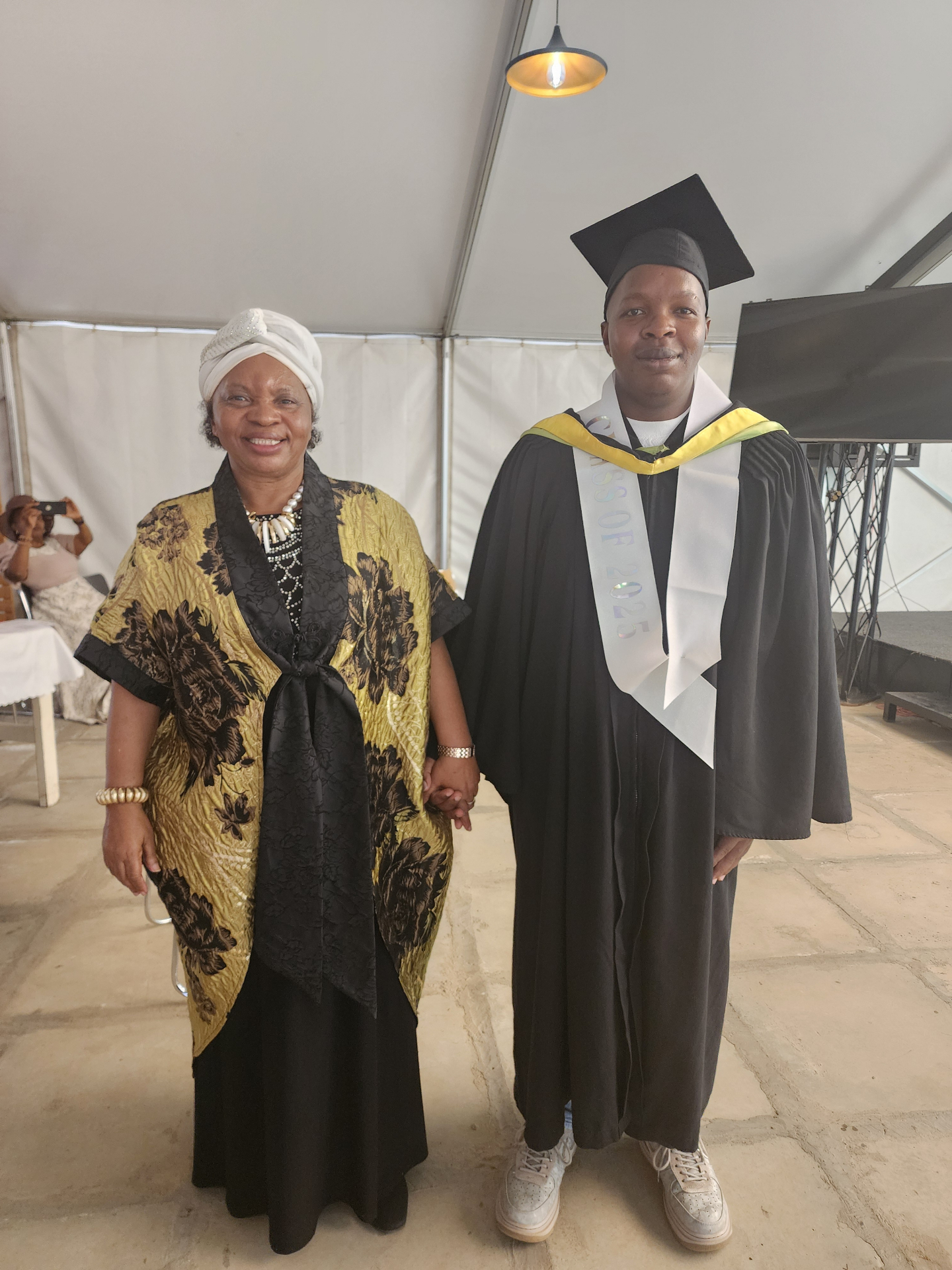 Samuel Mwangi in cap and gown on his graduation day at Jomo Kenyatta University, standing with Charisma Tumaini founder Dr. Grace Manjau